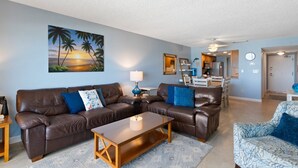 A living room with a brown leather sofa, a wooden coffee table, and a blue patterned armchair. There is a painting of a beach scene on the wall, a lamp, and a kitchen area in the background. - Prime Beachfront Balcony - Madeira Norte #311 (Madeira Beach)
