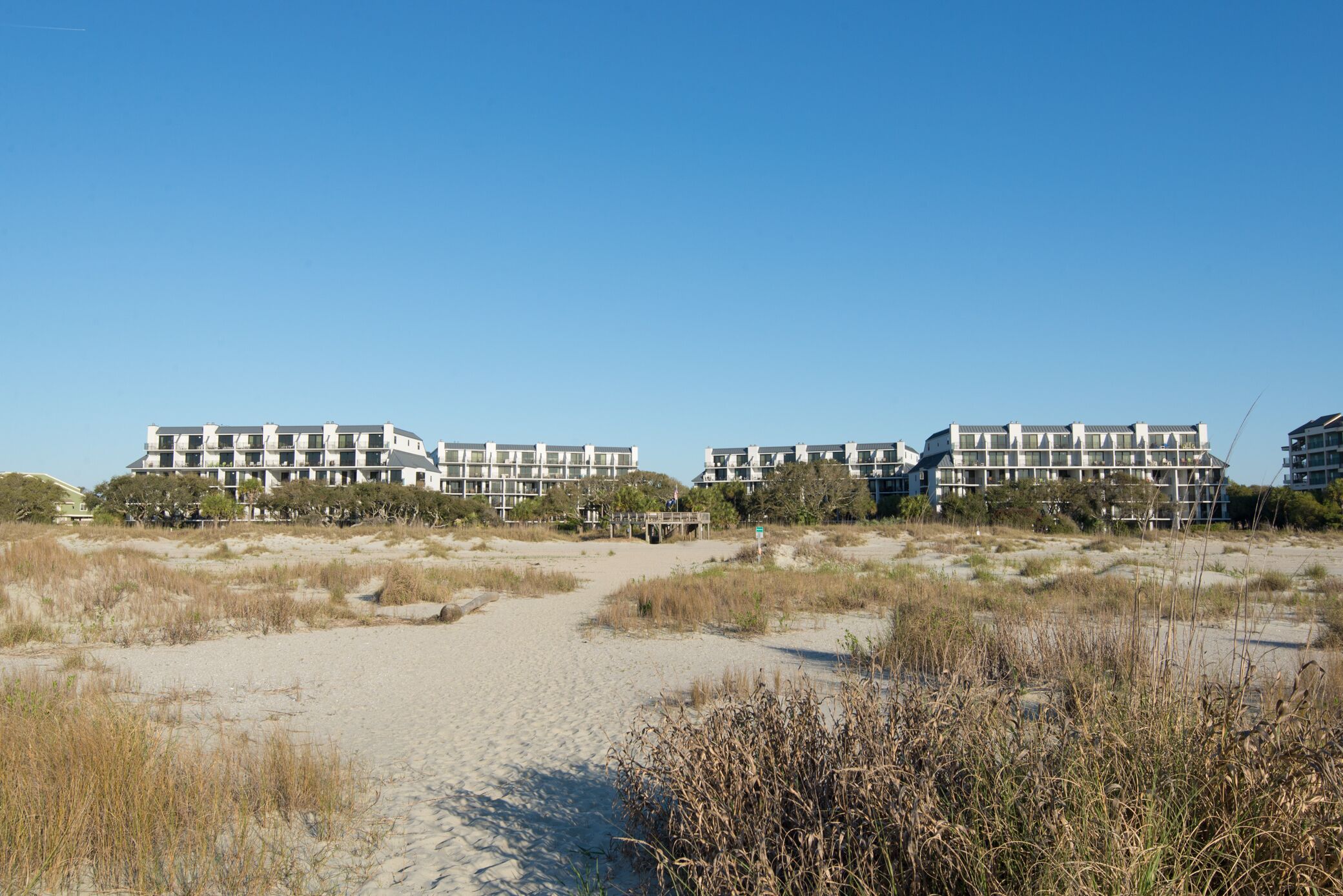 A beachfront hotel with multiple buildings, a sandy path leading to the beach, and surrounding dunes.