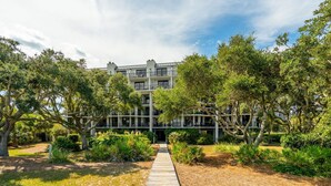 A multi-story building with a central courtyard, surrounded by lush greenery and a wooden pathway. - Shipwatch A204: Oceanfront condo, stunning views & private balcony retreat (Isle of Palms)