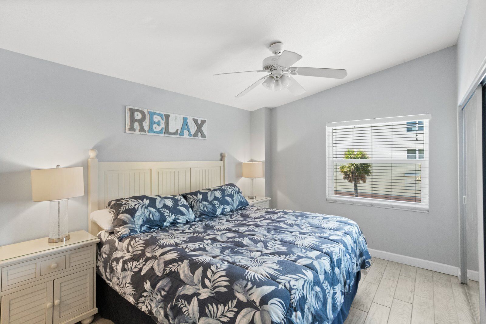 A bedroom with a bed, two bedside tables, a ceiling fan, a window with blinds, and a wall-mounted sign that says "RELAX".