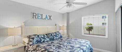 A bedroom with a bed, two bedside tables, a ceiling fan, a window with blinds, and a wall-mounted sign that says "RELAX".