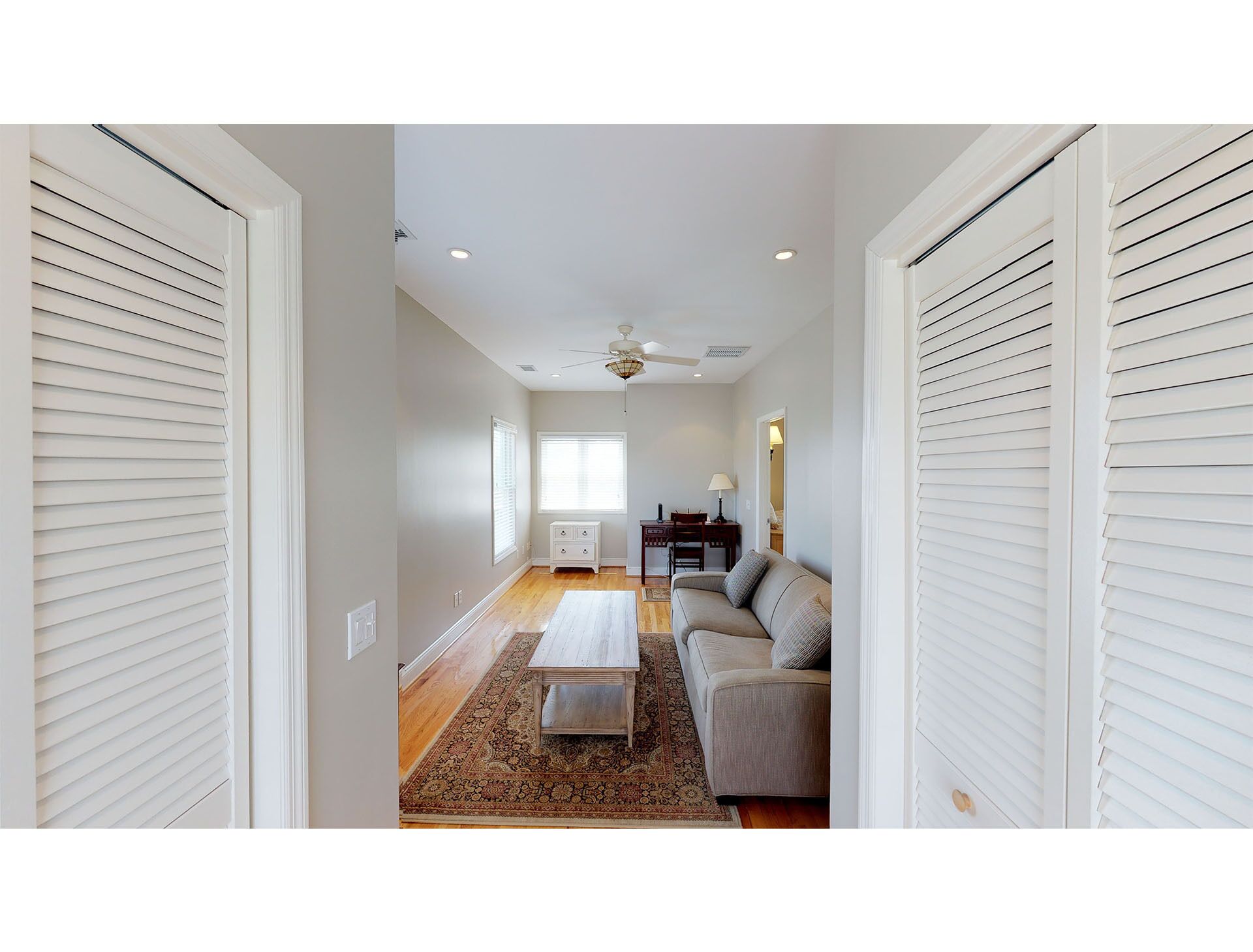 A spacious room with a ceiling fan, a wooden coffee table, a sofa, and a desk with a lamp.