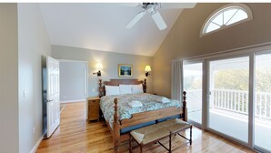 Ein Schlafzimmer mit einem hölzernen Bett, einer Bank, einem Deckenventilator, einem Nachttisch, einer Lampe und einem großen Fenster mit Balkon.