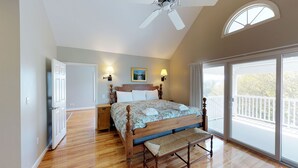 A bedroom with a wooden bed, a bench, a ceiling fan, a nightstand, a lamp, and a large window with a balcony.