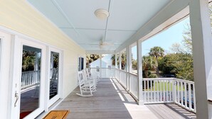 A covered porch with white railings, a ceiling fan, and a view of a blue house and palm trees in the background.