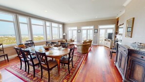A spacious living area with a large dining table, multiple seating options, and a view of the beach through large windows. - Three story home with sweeping ocean views and game room in Wild Dunes (Isle of Palms)