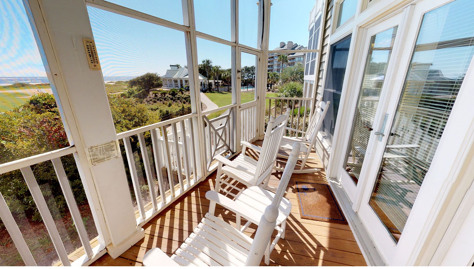 A wooden deck with white chairs and a glass balustrade overlooking a landscaped area and buildings.