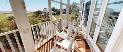 A wooden deck with white chairs and a glass balustrade overlooking a landscaped area and buildings.
