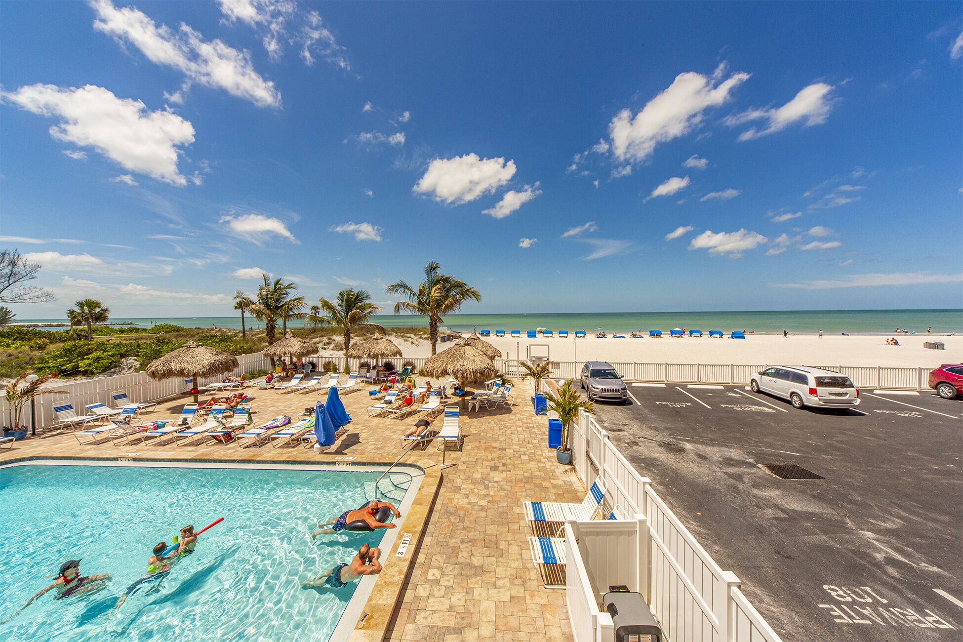 Un espace piscine avec des chaises longues et des parasols, un parking et une vue sur la plage et l’océan.