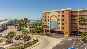 A beachfront hotel with a mural of waves and a sun on the facade, a parking lot with several cars, and a clear view of the beach and ocean. - Beach Front Balcony - Beach Place #107 (Madeira Beach)