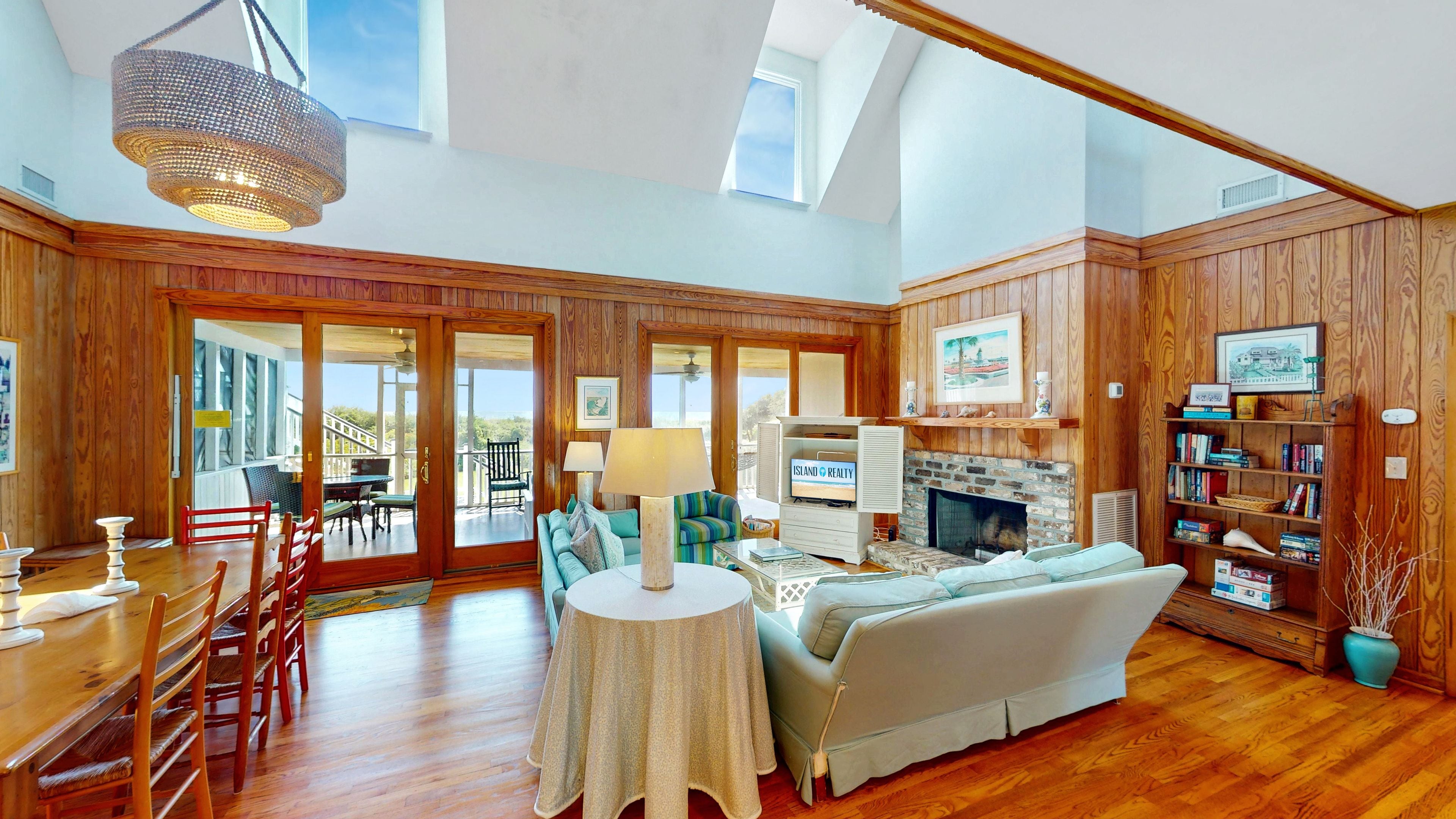 A spacious living room with a fireplace, wooden paneling, a dining area with a table and chairs, and a view through large glass doors.