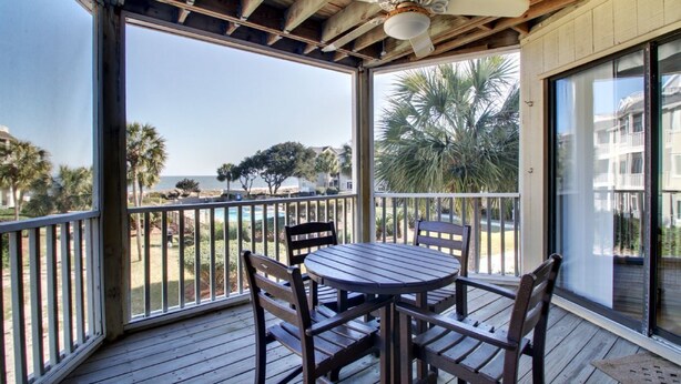 A covered outdoor area with a table and chairs, a ceiling fan, and a view of the ocean and palm trees. - Ocean View condo in Wild Dunes (Isle of Palms)