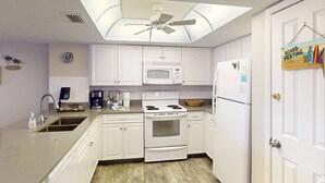 Eine moderne Küche mit weißen Schränken, einer Mikrowelle, einem Herd und einem Kühlschrank. Es gibt einen Deckenventilator, eine Uhr und ein Willkommensschild an der Wand.