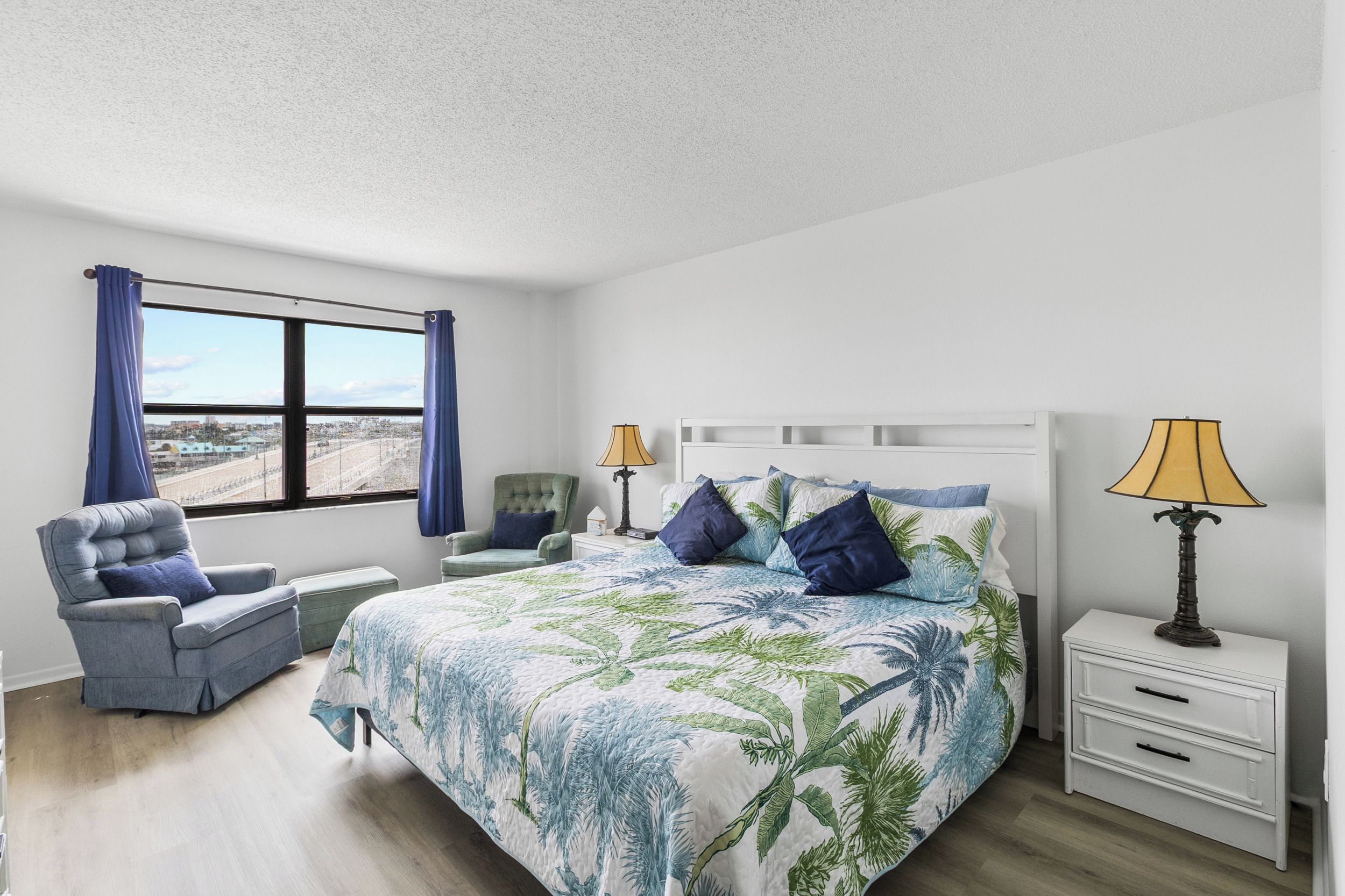 Un dormitorio con cama, una silla, una mesita de noche, una lámpara y una ventana con cortinas.