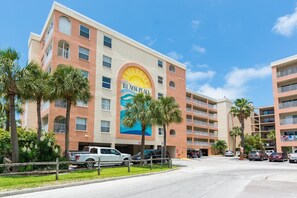 Un hotel de varios pisos con un llamativo mural de sol y olas en la fachada, palmeras y un estacionamiento con varios vehículos.