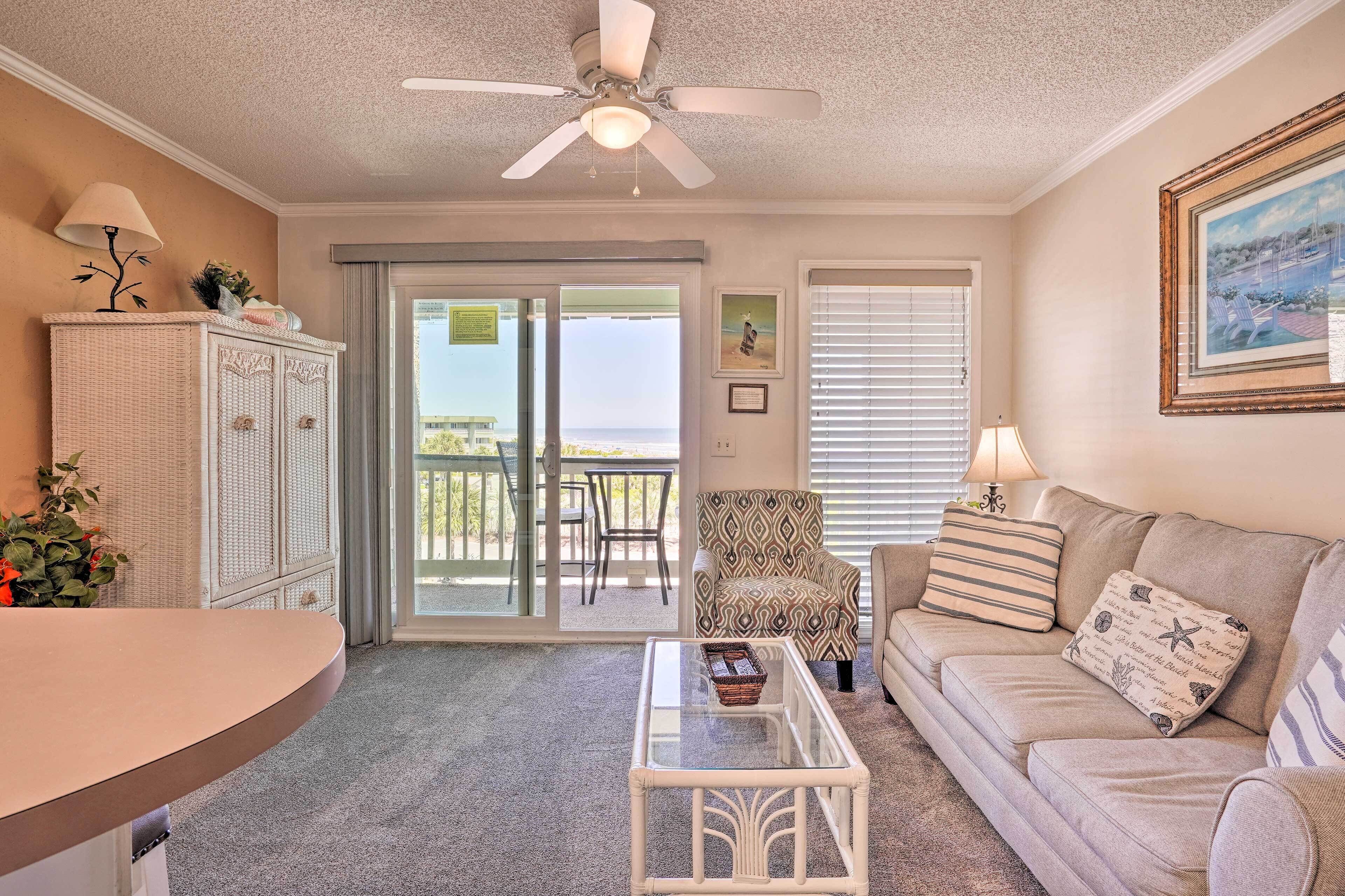 A living room with a sofa, armchair, and a glass coffee table. There is a ceiling fan, a wicker cabinet, and a view through the glass door to the outside.
