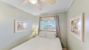 Un dormitorio con cama, ventilador de techo, ventana con cortinas y dos cuadros en las paredes.