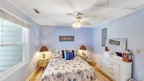 Une chambre à coucher comprenant un lit, deux tables de chevet avec des lampes, un ventilateur de plafond, une commode et une fenêtre avec des stores.