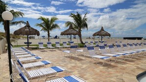 A beachside lounge area with rows of sun loungers and thatched umbrellas, a pool, and palm trees.