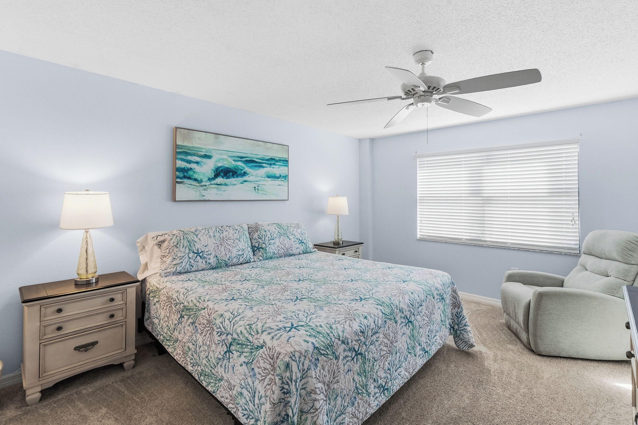 A bedroom with a bed, two bedside tables, a ceiling fan, a window with blinds, and a chair.