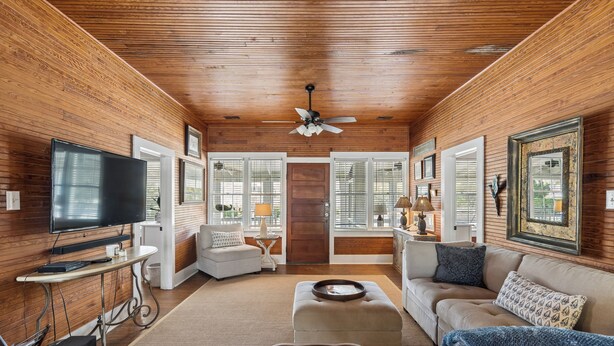 A living room with wooden walls, a ceiling fan, a television, a sofa, a coffee table, and a door with windows.