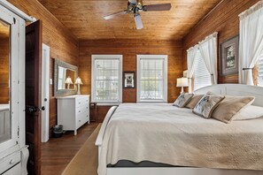 Ein Schlafzimmer mit Holzbalkendecke, einem Bett mit weißer Bettdecke, zwei Nachttischlampen, einem Nachttisch, einem Schminktisch und einem Spiegel.