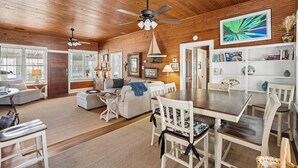 A spacious living room with a wooden ceiling, a dining area with white chairs, and a bookshelf with books and decorative items.