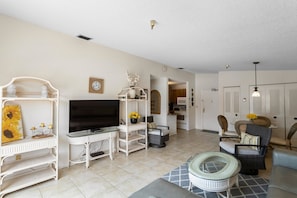 A living room with a TV, white shelving, a dining area, and a kitchen in the background.