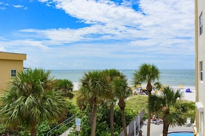 A beach view from a balcony with palm trees, a pool, and a building with a sign. - Beach View - John's Pass - Madeira Norte #208 (Madeira Beach)