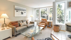 Ein Wohnzimmer mit grauem Sofa, einem Glastisch, einem Kamin und großen Fenstern mit Blick auf Bäume.