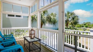 A covered porch with a table and chairs, a birdcage, and a view of a landscaped area with palm trees.