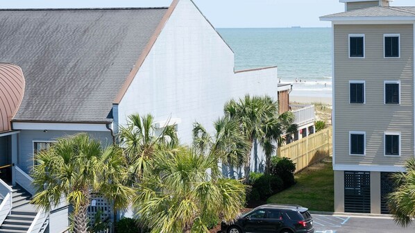 A coastal residential area with a white building, a black car, and a view of the ocean. - Pool, Rooftop Ocean view, beach/shops nearby! (Isle of Palms)