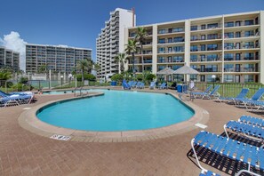 Un espace piscine d’hôtel avec une piscine à l’eau d’un bleu limpide, des chaises longues et, au fond, un bâtiment doté de nombreux balcons.
