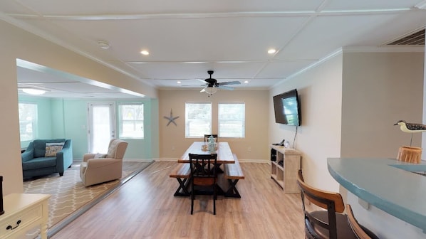 A spacious living area with a wooden floor, a dining table with benches, a ceiling fan, a TV mounted on the wall, and a starfish decoration.