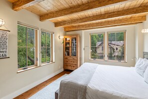 Une chambre avec un grand lit, des poutres apparentes en bois au plafond et deux fenêtres donnant sur des arbres et une maison.