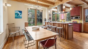 A modern kitchen with wooden beams, a dining area with a large table and chairs, and a view of the outdoors through large windows.