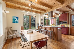 A modern kitchen with wooden beams, a dining area with a large table and chairs, and a view of the outdoors through large windows.