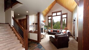 A spacious living area with a stone fireplace, a large window offering a view of trees, and a wooden staircase. - Home Across from Purg - Views, Deck/Fire Pit/Ping Pong/Shuffleboard (Durango)