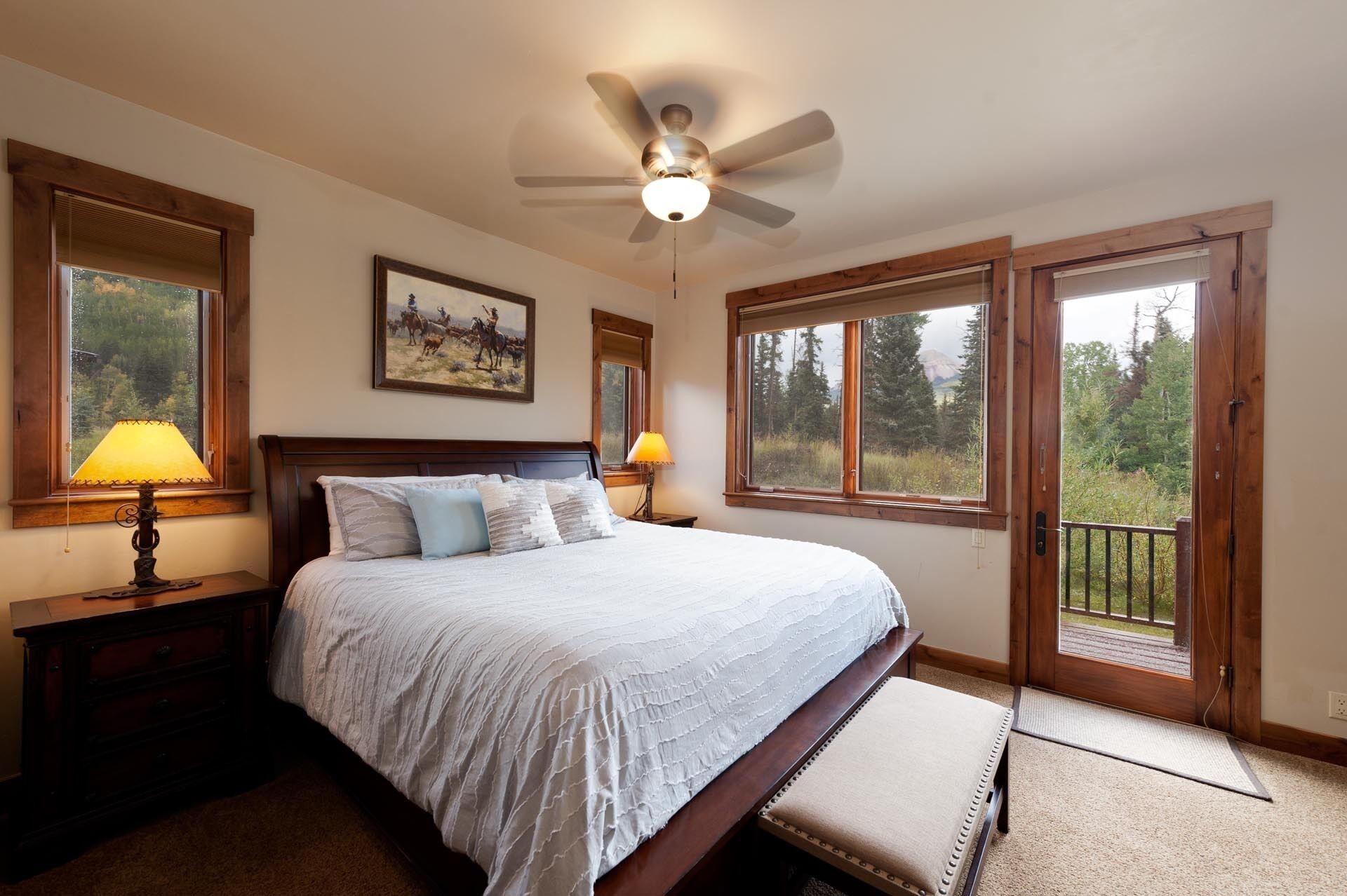 A bedroom with a large bed, two bedside lamps, a ceiling fan, and a view of the outdoors through a large window.