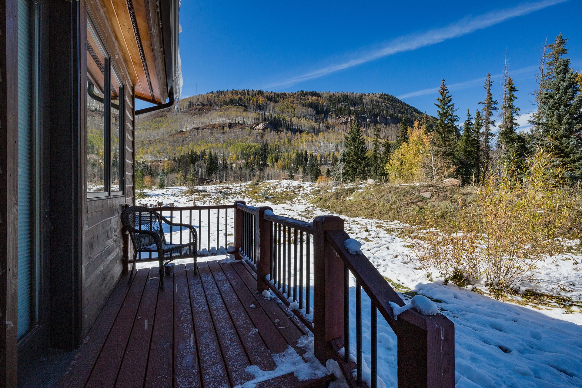A wooden deck with a metal railing, overlooking a snowy landscape with trees and a mountain in the background.