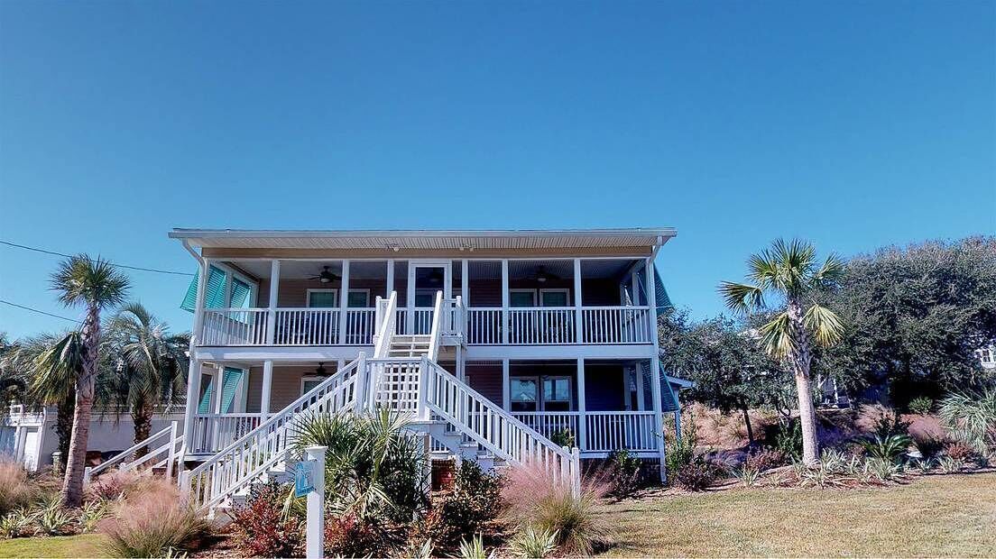 A two-story building with a balcony on the upper floor, surrounded by palm trees and shrubs.