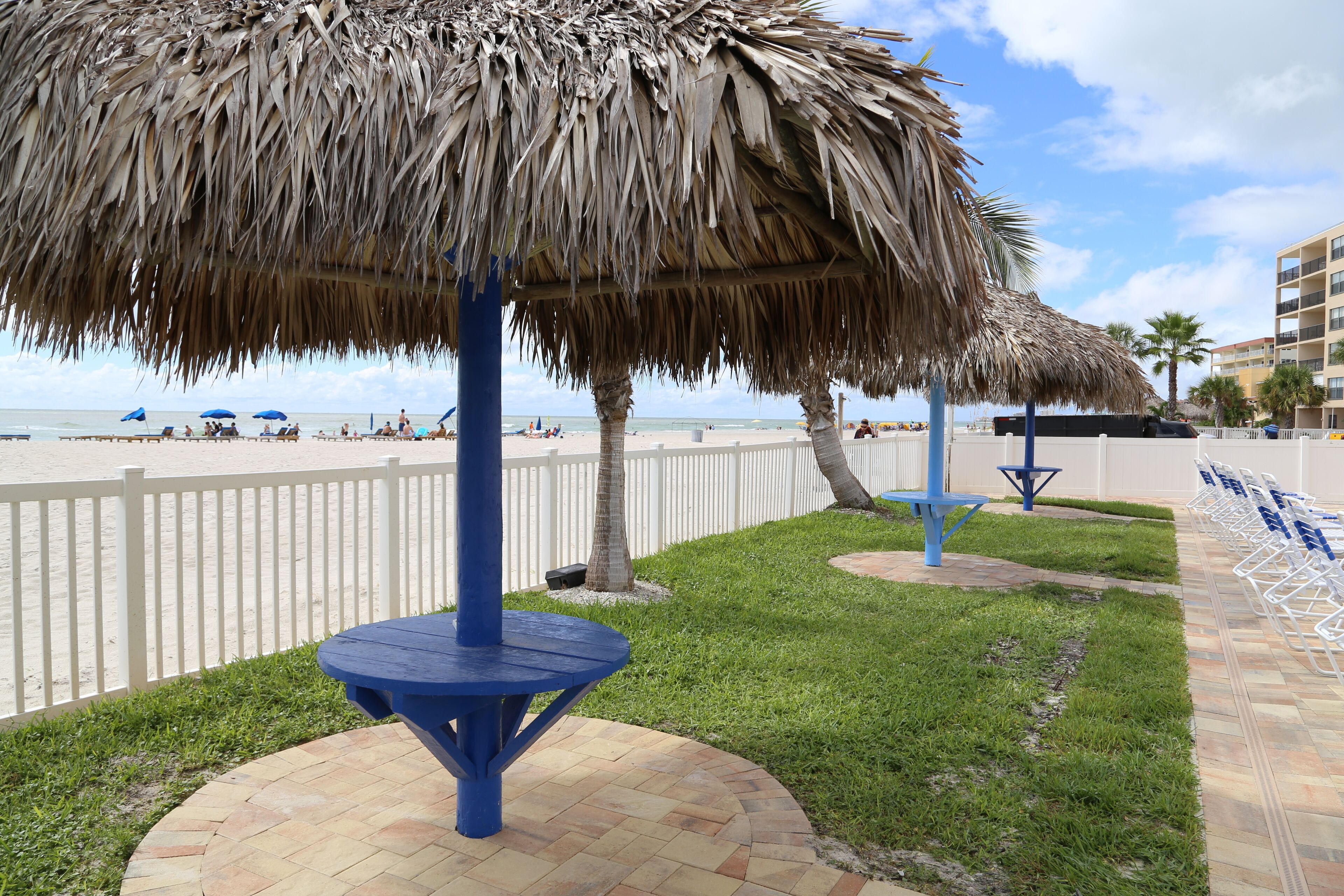 Un espace aménagé sur la plage, avec un parasol en chaume, une table bleue et une clôture blanche.