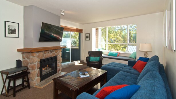 A living room with a stone fireplace, a large blue sofa, a coffee table, and a television mounted on the wall. - Eagle Lodge 432 MOUNTAIN VIEWS + VILLAGE LOCATION! Professionally Managed + Cleaned (Whistler)