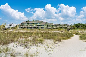 A beachfront property with multiple green-roofed buildings, a sandy pathway leading to the beach, and a clear blue sky.