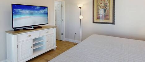 A bedroom with a bed, a TV on a dresser, a painting on the wall, and a floor lamp.