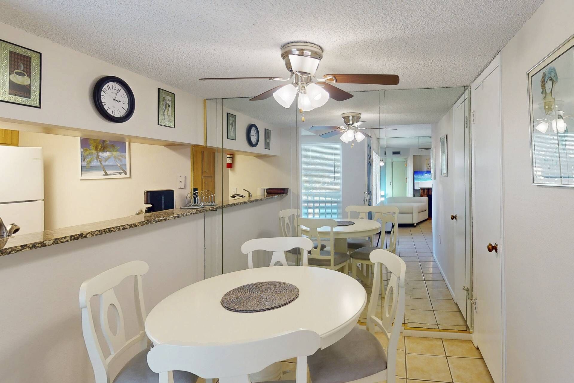 Un comedor con una mesa y sillas blancas, un ventilador de techo y una zona de cocina con un refrigerador y un reloj.