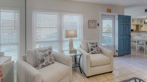 A living room with two sofas, a table, and a lamp. There is a door leading to a kitchen and a balcony. - 2-Level Madeira Beach Condo, Pool, Marina, Walk to Beach, Shops,  Dining +Tickets to Top Activities! (Madeira Beach)