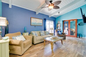 A living room with a blue wall, wooden flooring, a beige sofa, a glass coffee table, and a ceiling fan.