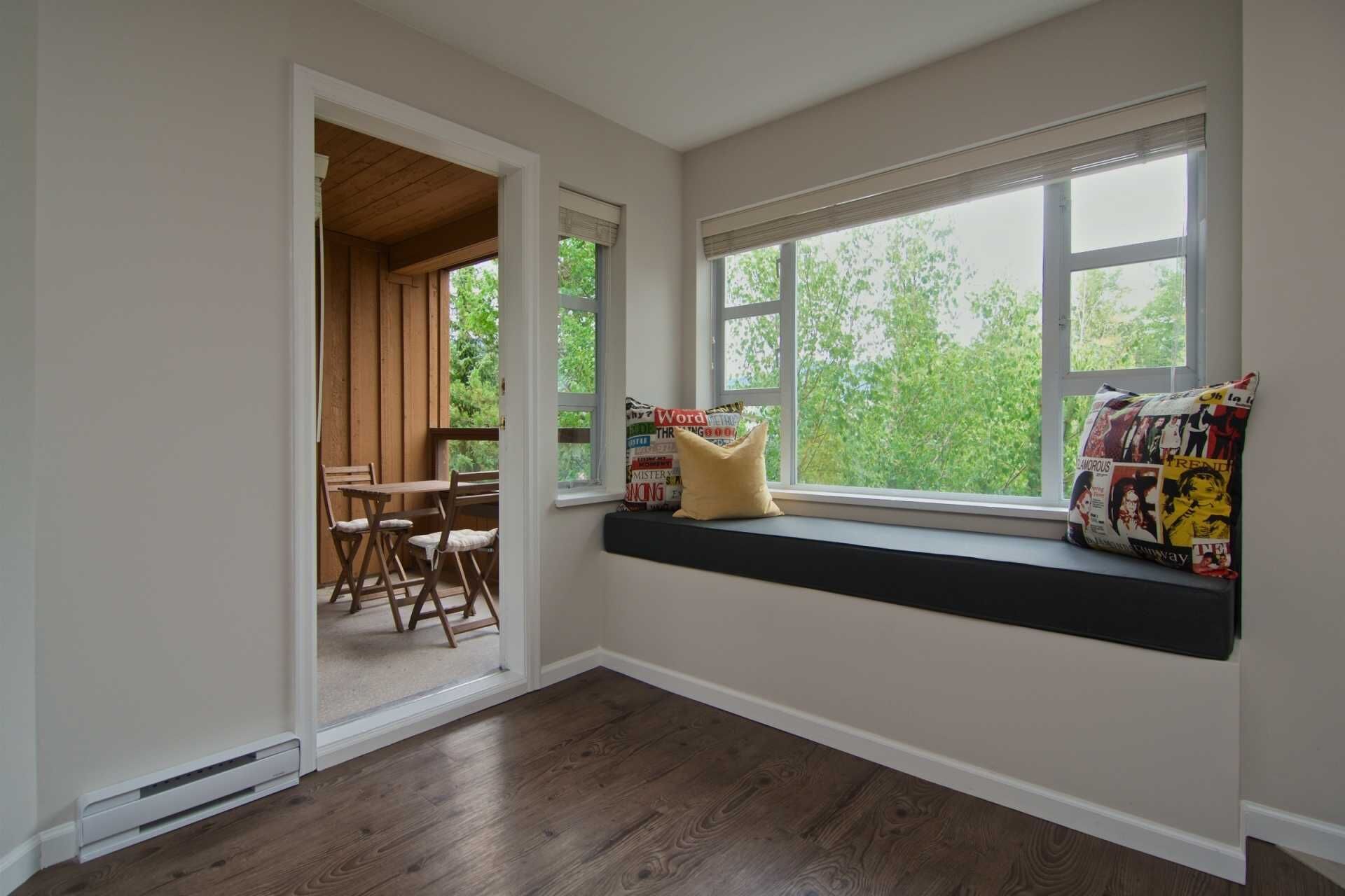 A room with a built-in bench under a large window, a wooden door leading to another room, and a small table with chairs.