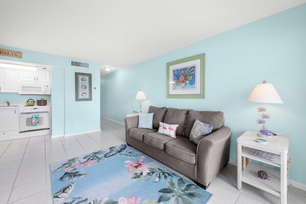 A modern living room with a grey sofa, a floral rug, and a kitchen area with white cabinets and a microwave.